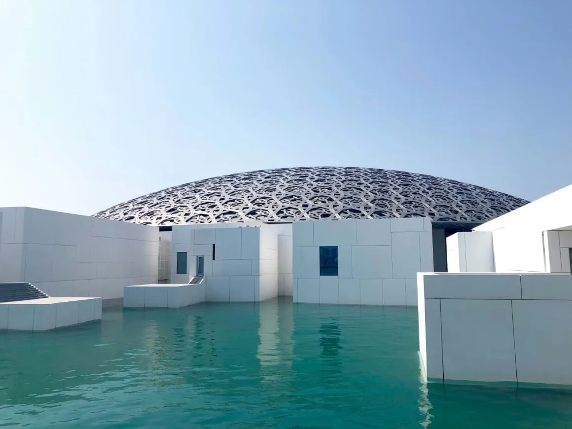 The Louvre Abu Dhabi on Saadiyat Island. a white building with a metal sculptural roof over blue water