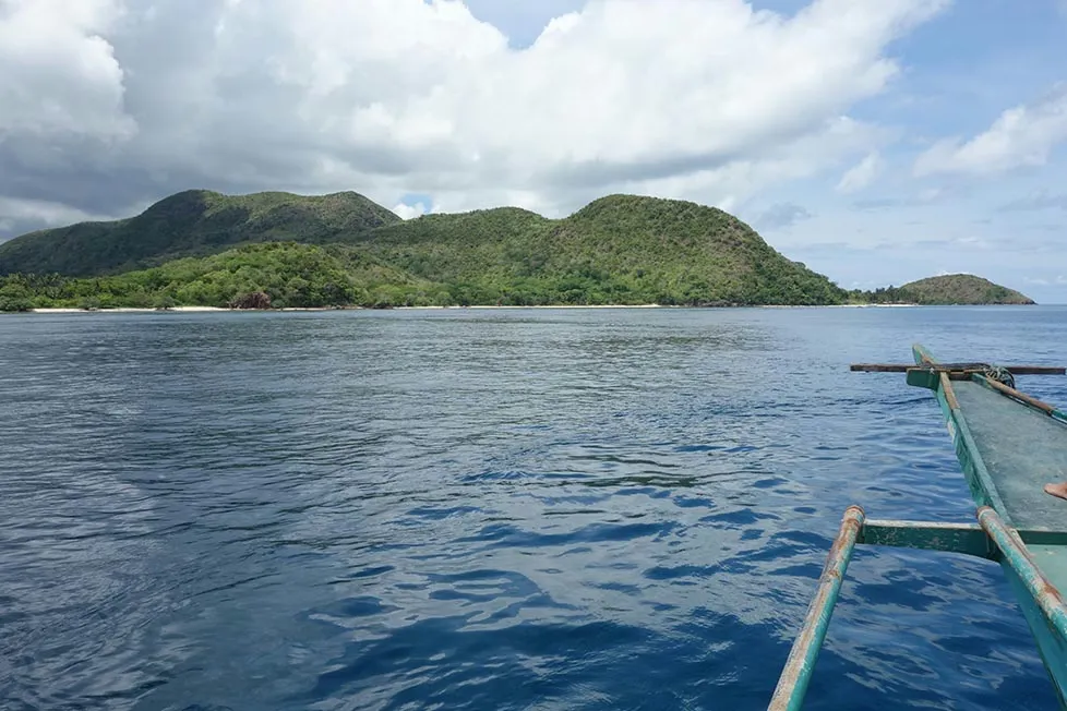 The front of a traditional wooden boat as it cuts through the deep blue sea headed for a jungle covered island.