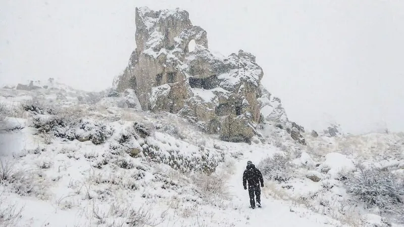 A person walking/ hiking on a mountain towards a rock formation in Cappadocia, Turkey. The landscape is covered in snow and it is snowing heavily still.