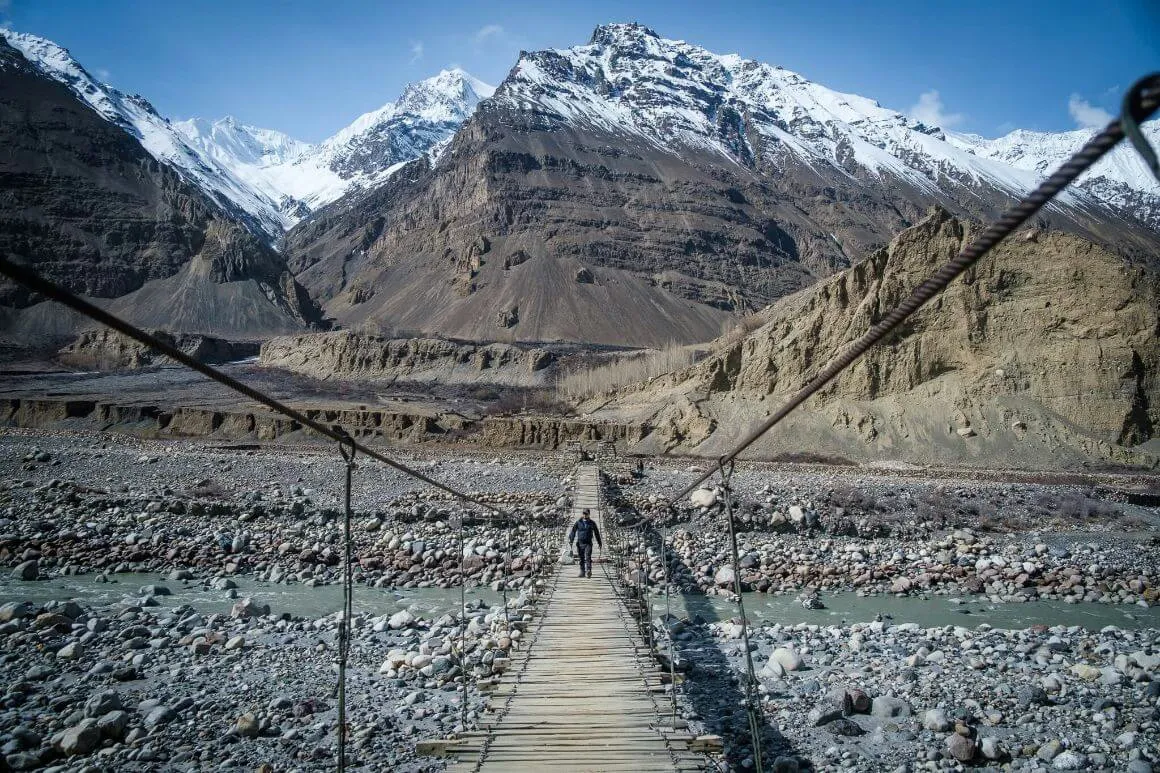 Will walking across a wooden swing bridge that goes over a river in a valley surrounded by massive snow covered mountains