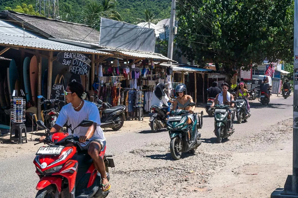 backpackers and surfers riding on scooters past clothing and surfboard rental shops in Kuta Lombok on a sunny day